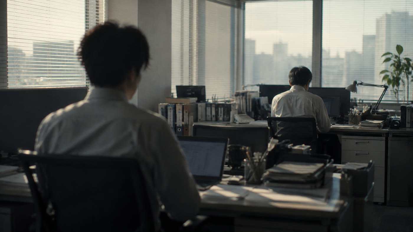 オフィスで、若い社員の視線の先に座る年上の社員の背中